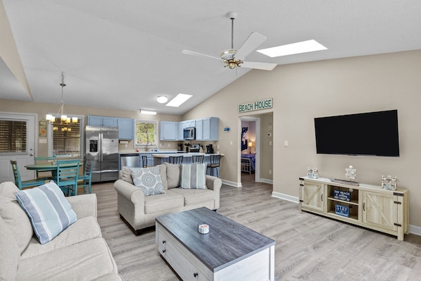 A living room with a sofa, coffee table, and TV. There's a kitchen area with a fridge, microwave, and cabinets. A sign on the wall reads "BEACH HOUSE".