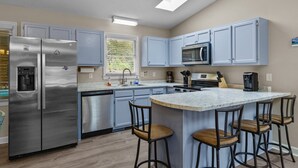 Una cocina moderna con electrodomésticos de acero inoxidable, armarios azul claro, una isla central con taburetes y una ventana con persianas.
