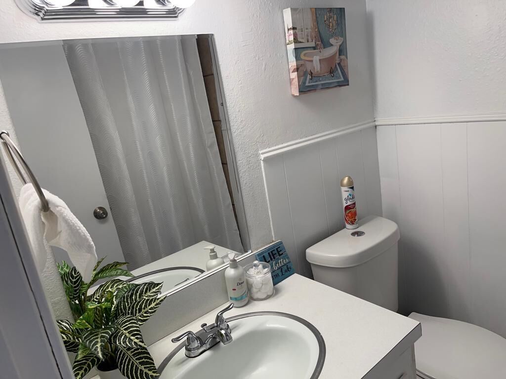 Un baño con lavamanos blanco, espejo y inodoro. Hay una planta a la izquierda y un cuadro sobre el inodoro.