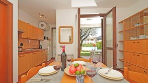 Ein Essbereich mit Tisch für vier Personen, eine Küche mit Holdelementen und Blick auf einen Garten durch eine offene Tür.