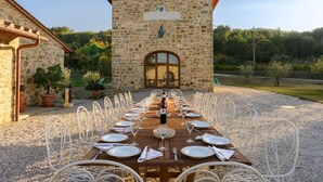 Un edificio de piedra con una mesa de madera dispuesta para comer, rodeada de sillas blancas.