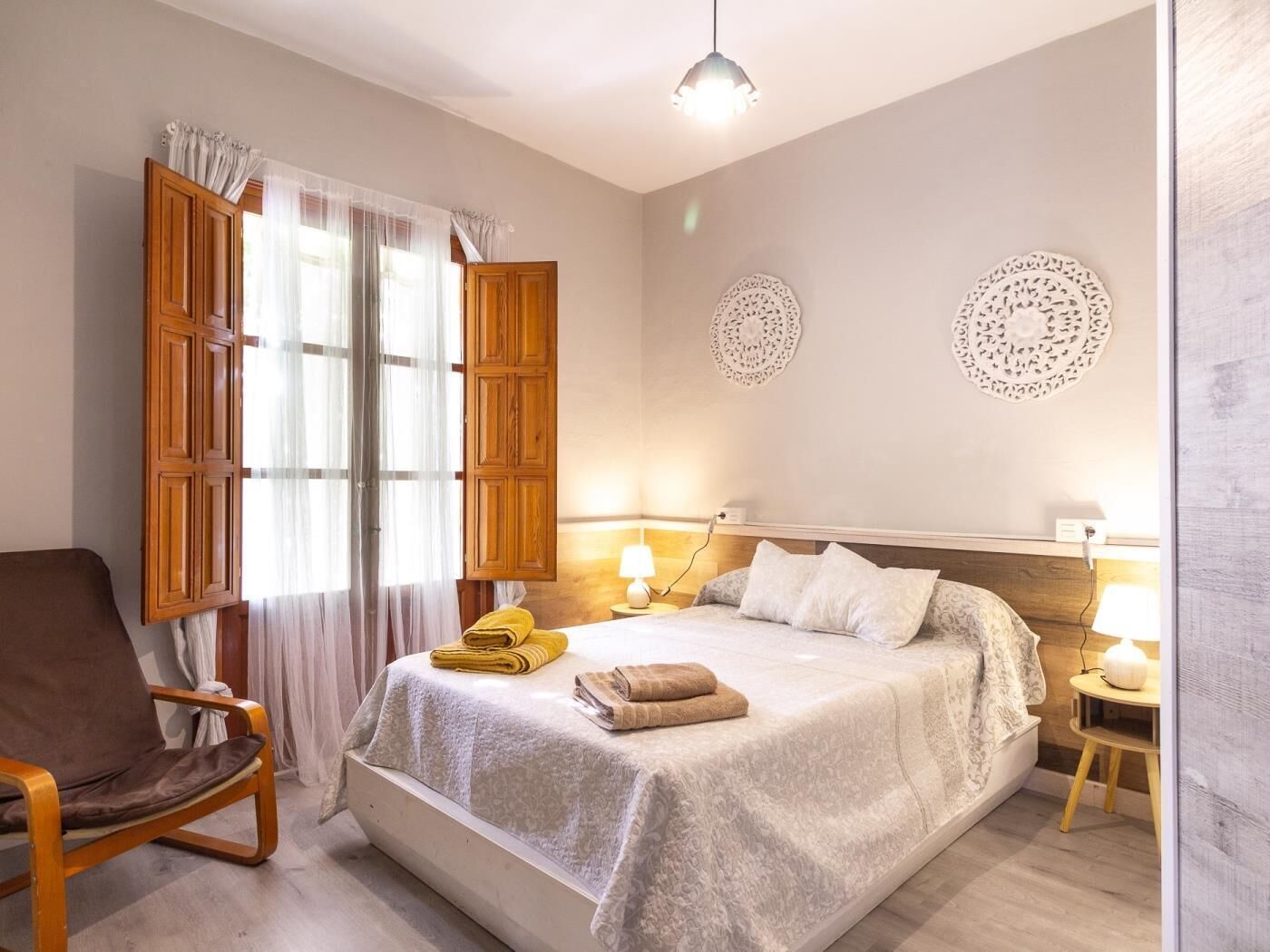 Een hotelkamer met een bed, een stoel, een nachtkastje met een lamp, en een raam met gordijnen.