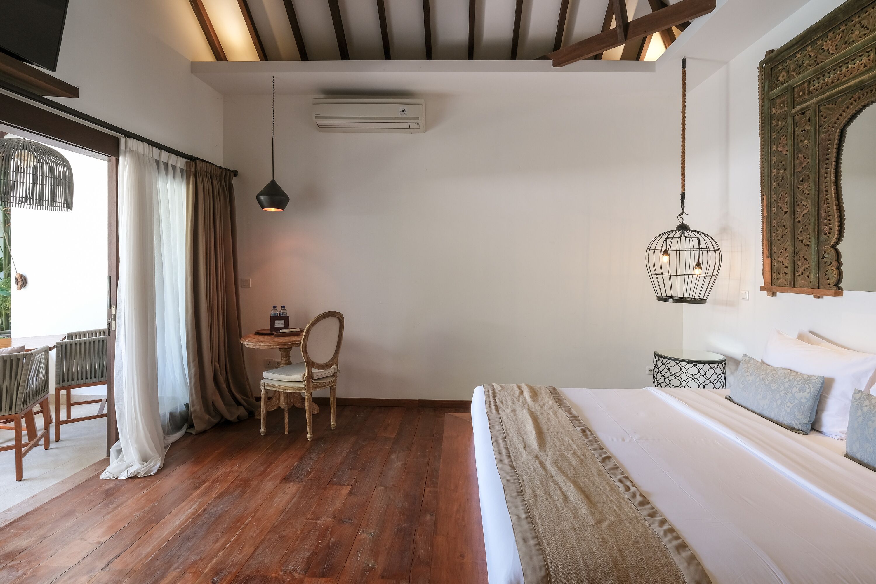 A bedroom with a wooden floor, a bed with white bedding, a desk with a chair, and a hanging light fixture.