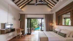 A bedroom with a large bed, a TV, a dining area with a table and chairs, and a view of an outdoor swimming pool. - Aram Residence (Canggu)