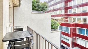 Parveke, jossa on pöytä ja tuoleja, näkymä punaiseen rakennukseen, jossa on useita ikkunoita, sekä pieni puutarha-alue.