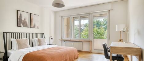 A bedroom with a bed, a desk, a chair, a window, and artwork on the wall.