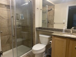 A modern bathroom with a glass-enclosed shower, a toilet, and a granite countertop with a sink and mirror. - Sea Colony 2 BR, Screened Porch, Steps to Pool, Pickleball, Tennis (Bethany Beach)