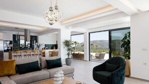 A spacious living area with a sofa, a coffee table, and a dark armchair. There is a dining area with a table and chairs, and a kitchen in the background. - Villa Empire Ultimate Luxury villa with heated pool (Kefalas)
