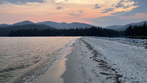 A beach with snow on the right side, a body of water in the middle, and mountains in the background.