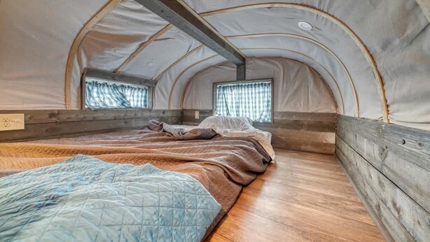Een tent met een houten vloer, twee ramen met gordijnen, en een bed met een blauwe deken.