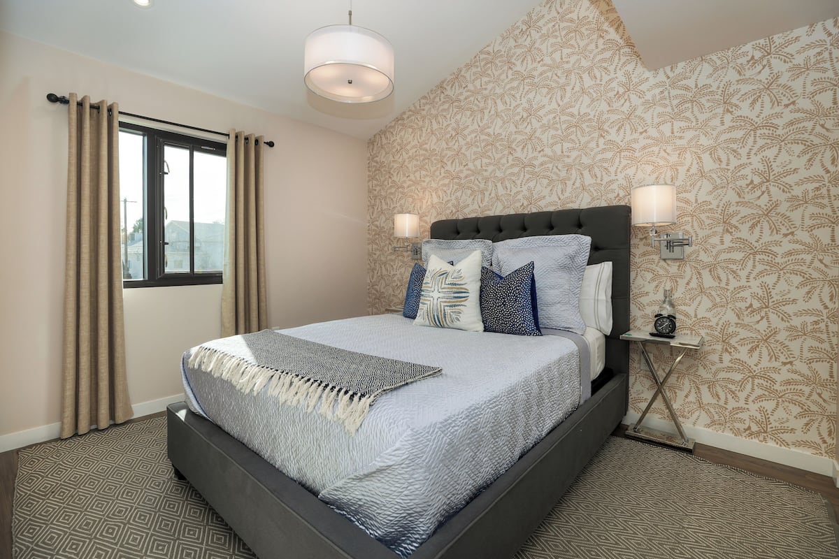 A bedroom with a bed, curtains, a patterned wall, and a nightstand with a lamp.