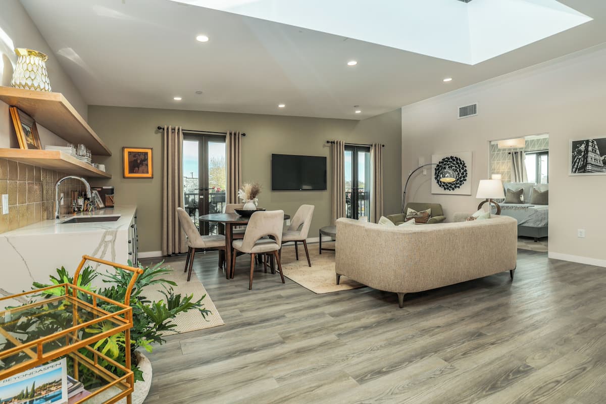 A spacious living area with a high ceiling, a dining table with chairs, a sofa, and a TV mounted on the wall.