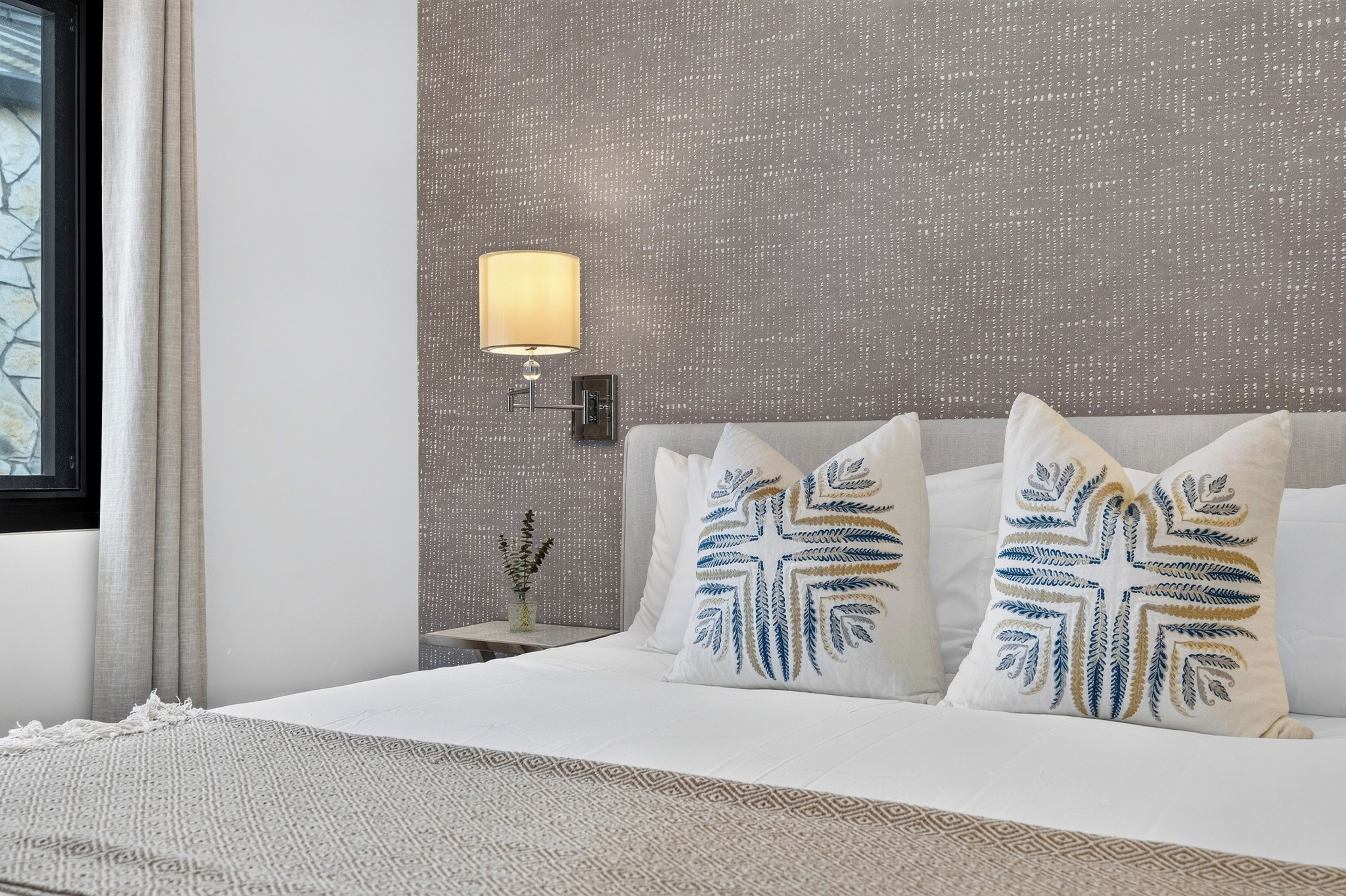 Ein Hotelzimmer mit einem Bett, Kissen, einem Nachttisch, einer Lampe und einem fenster mit Vorhängen.