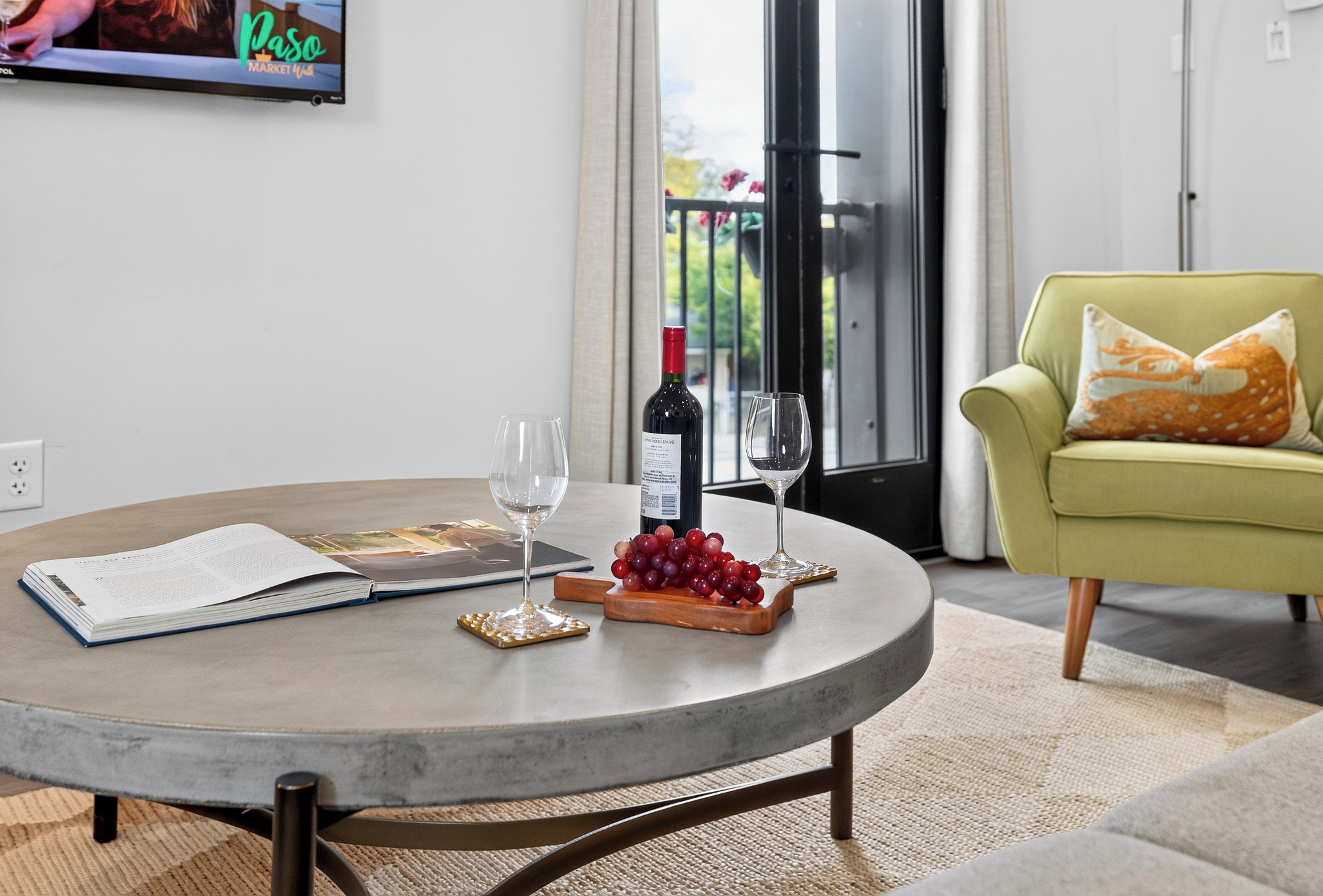 Una mesa redonda con una botella de vino, dos copas, un libro y una bandeja de frutas en un moderno salón con un sillón verde y un televisor mostrando 'Paso Market 4th'.