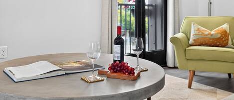 A round table with a wine bottle, two glasses, a book, and a fruit tray in a modern living room with a green armchair and a TV displaying 'Paso Market 4th'.