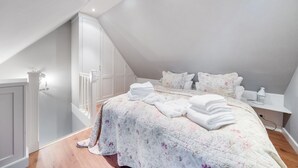 Een slaapkamer met een schuine plafond, een bed met bloemenbedrukking, een nachtkastje met een lamp en een witte kledingkast.