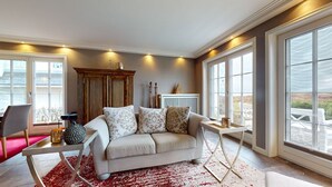Een ruime woonkamer met een beige bank, een houten kast en een witte tafel. Grote ramen bieden uitzicht op het omliggende landschap.