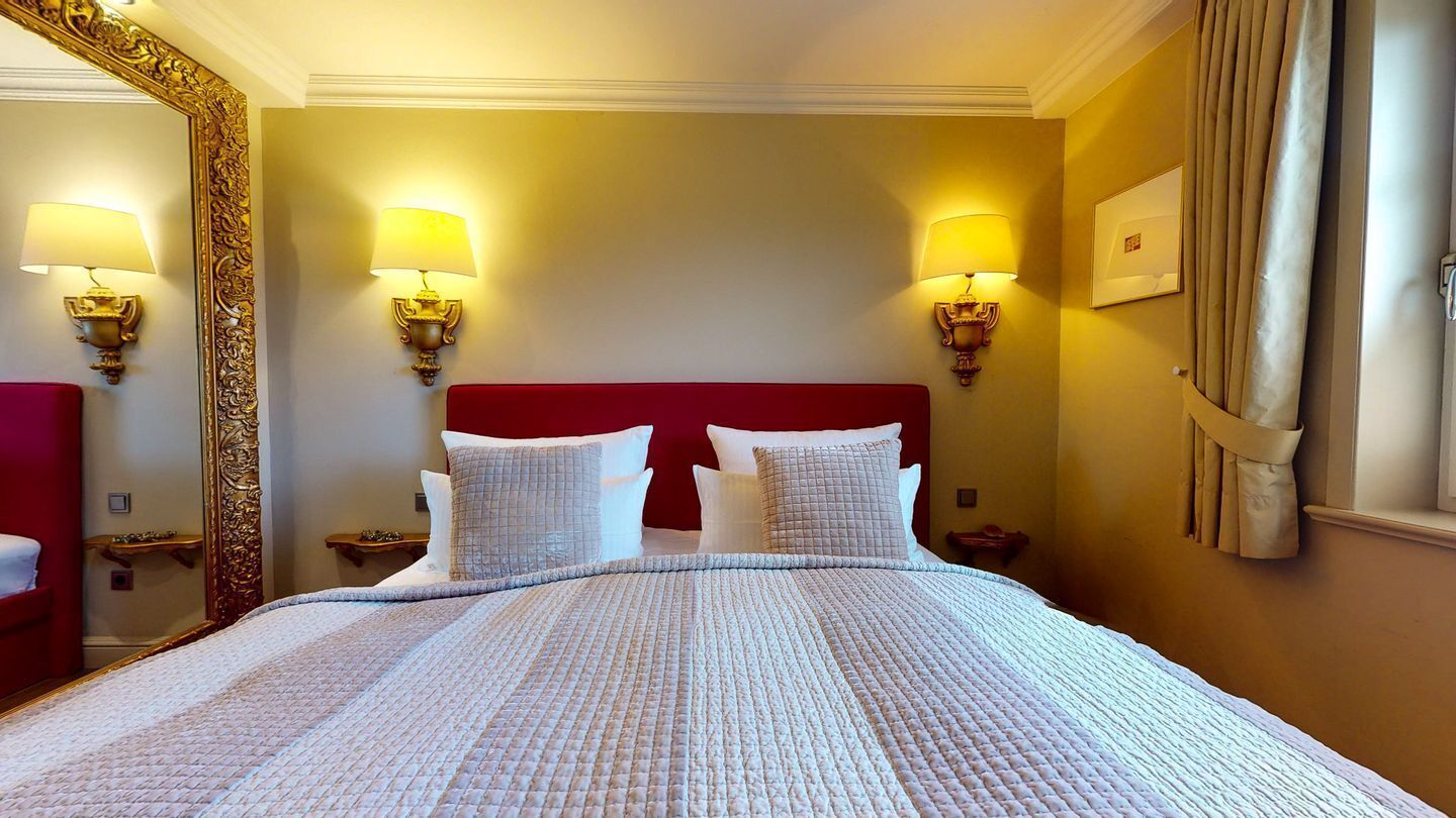A hotel room with a large bed, red headboard, and two wall-mounted lamps.