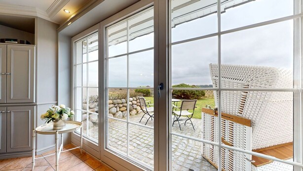 A room with a large window, a small table with a vase of flowers, and a wicker chair outside. - Pure Sylt - Exclusive luxury apartment in a quiet location on the Munkmarsch Wadden Sea. (Sylt)
