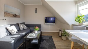 A modern living room with a grey sofa, a coffee table with a vase of flowers, a TV mounted on the wall, and a dining area with a table and chairs. - Sylt Like - Modernes Appartement in Zentraler Lage für bis zu Zwei Personen (Sylt)