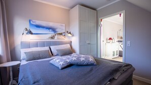 A bedroom with a bed, a nightstand, a wall-mounted lamp, a ceiling light, and a door leading to a bathroom. - Sylt Like - Modernes Appartement in Zentraler Lage für bis zu Zwei Personen (Sylt)