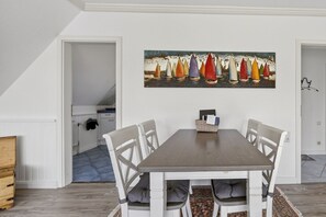 A dining area with a table and chairs, a painting above the table, and a kitchen in the background.