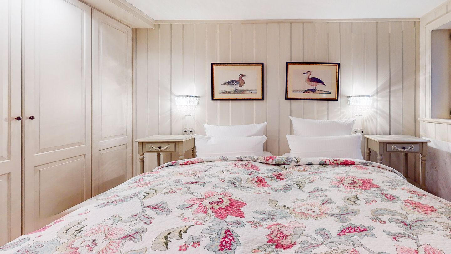 Une chambre à coucher avec une couvre-lit à motifs floraux, deux tableaux encadrés accrochés au mur, des lampes de chevet et une armoire en bois.