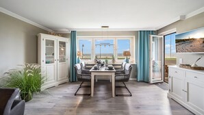Uma sala de jantar com mesa posta para quatro pessoas, rodeada por cadeiras de vime, um armário branco com portas de vidro e vista para a praia através de grandes janelas.