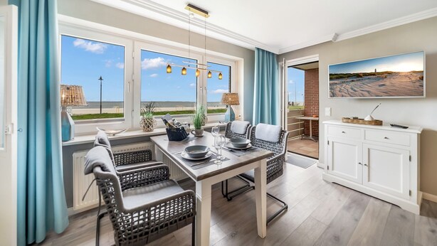 Uma área de refeições com vista para a praia, cadeiras de vime, uma mesa com pratos e copos, e uma televisão na parede.