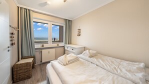Quarto de hotel com cama, uma mesinha, janela com cortinas, radiador e vista para a praia.