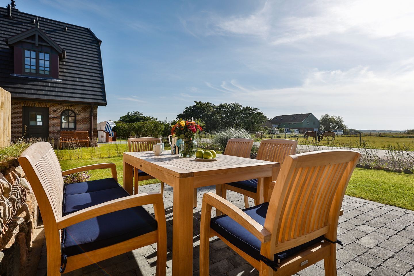 An outdoor wooden table and chairs set on a paved patio with a brick house in the background.
