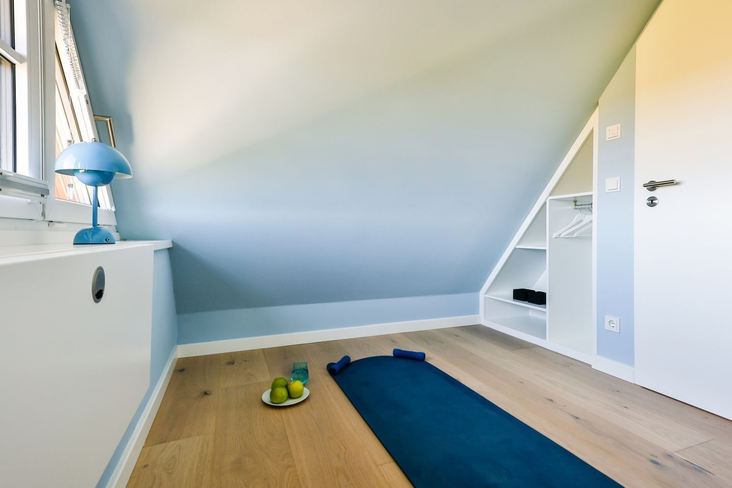 A room with a sloped ceiling, a blue floor mat, a small table with a blue lamp, and a shelf with a few items.