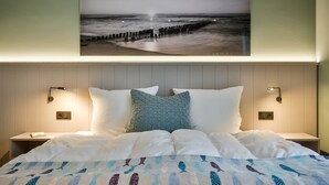 A bedroom with a bed, pillows, a wall-mounted picture of a beach, and bedside lamps. - Newly built house with garden and two terraces in a salt marsh location in Tinnum. - (Sylt)