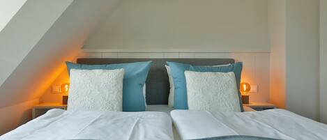 Une chambre avec un plafond en pente, un lit avec une literie blanche et bleue, deux tables de chevet avec des lampes et une tête de lit avec un panneau gris.