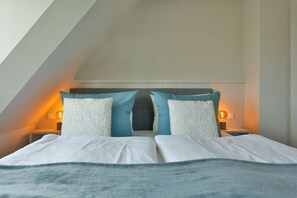 A bedroom with a sloped ceiling, a bed with white and blue bedding, two nightstands with lamps, and a headboard with a gray panel. - Exclusive new-build house for 4 people in a popular salt marsh location in Tinnum. (Sylt)