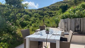 A wooden deck with a table and chairs, surrounded by lush greenery and a hill in the background. - Modern apartment for two people in a location absolutely close to the beach - House (Hörnum (Sylt))