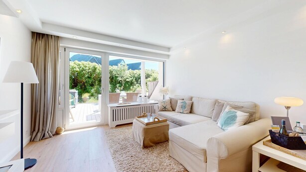 A modern living room with a sofa, a coffee table, and a lamp. There is a view of greenery outside through large windows. - Modern apartment for two people in a location absolutely close to the beach - House (Hörnum (Sylt))