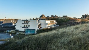 A modern building with a flat roof and large windows, surrounded by grassy terrain and traditional houses in the background. - Modern apartment for two people in a location absolutely close to the beach - House (Hörnum (Sylt))
