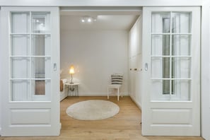 Una habitación con una puerta blanca grande, un tapete redondo, una mesita de noche con una lámpara y una silla.