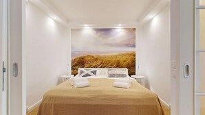 A hotel room with a bed, two bedside tables, a large landscape painting on the wall, and a door on the left. - Modern apartment for two people in a location absolutely close to the beach - House (Hörnum (Sylt))