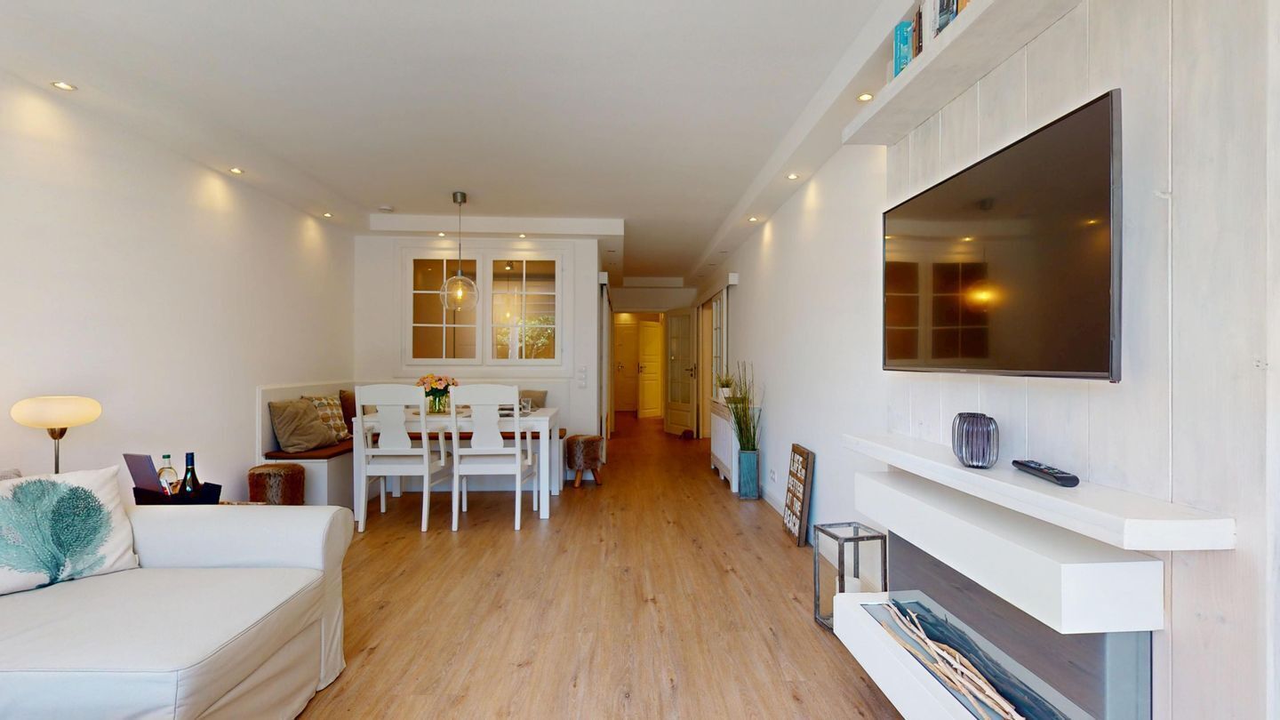 Una sala de estar moderna con un sofá blanco, una mesa de comedor con sillas, una chimenea y un televisor montado en la pared.