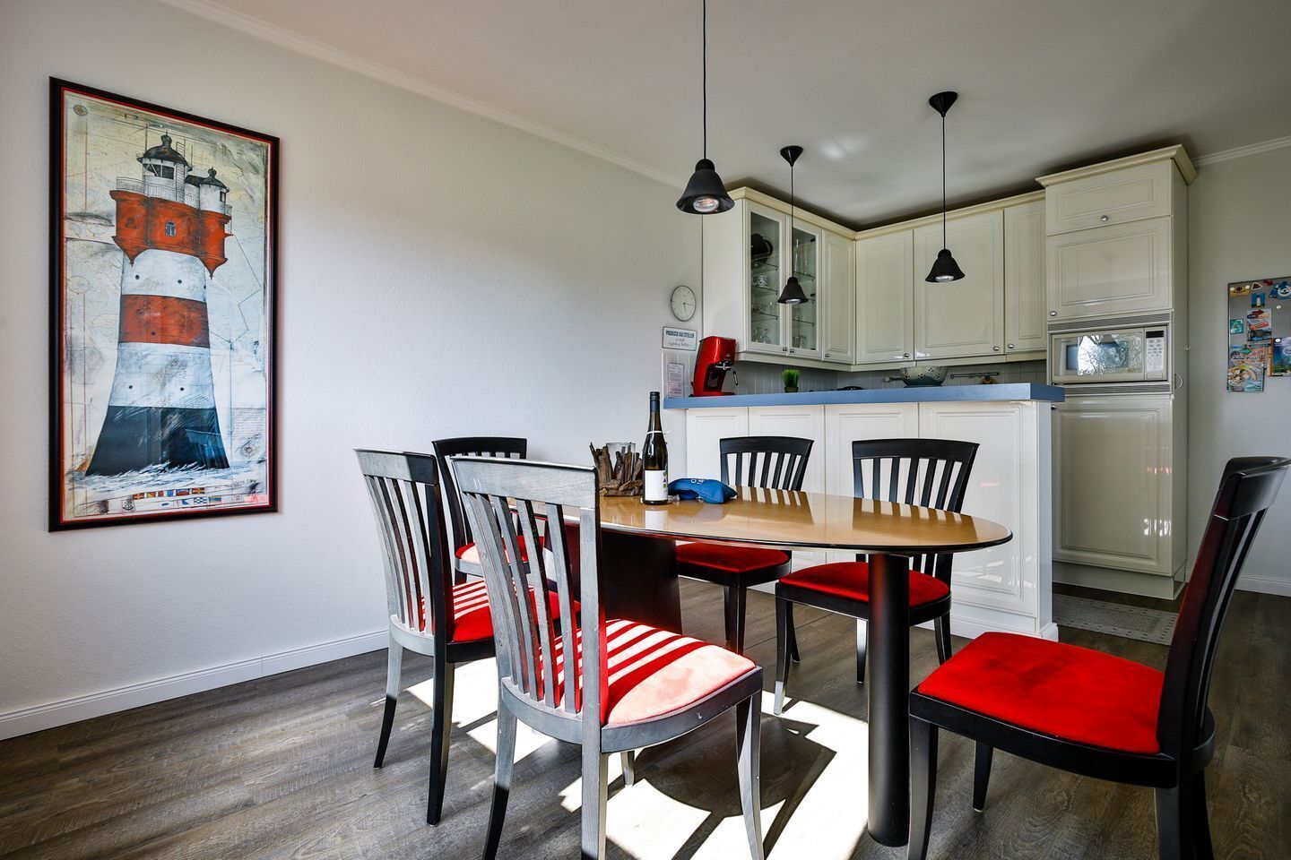Un coin repas avec une table et des chaises en bois, un tableau représentant un phare, et une cuisine avec des meubles blancs et un four à micro-ondes.