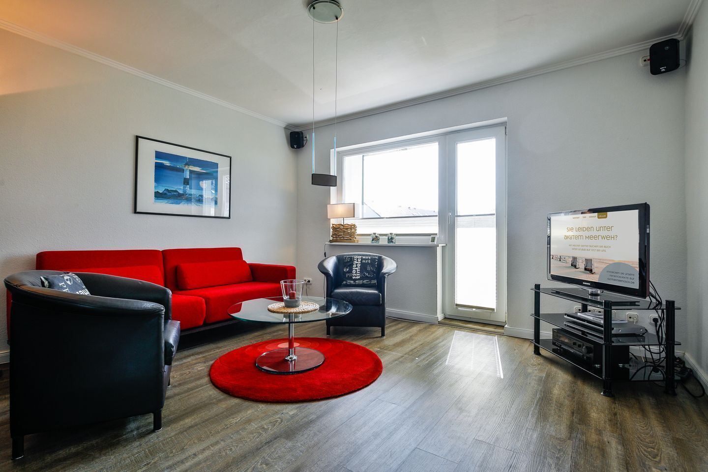 A modern living room with a red sofa, a black armchair, a glass coffee table, and a red rug. There is a large window with a view of the outside, a flat-screen TV, and a speaker mounted on the wall.