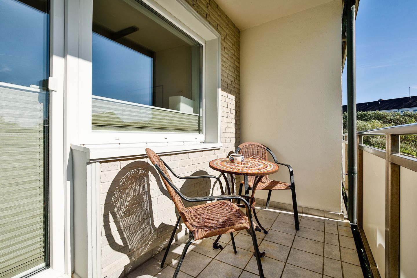 Un balcon avec deux fauteuils en osier et une petite table ronde, une fenêtre et une vue sur le ciel.