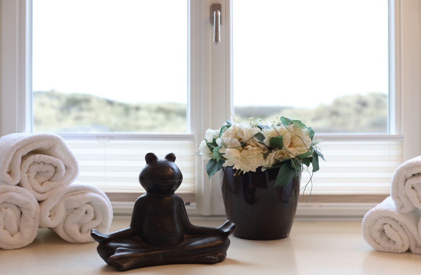 Una ventana con toallas enrolladas, un objeto con forma de oso oscuro y un jarrón con flores.