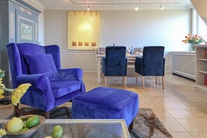 A living room with a blue sofa, matching ottoman, and two matching chairs. A dining table with chairs is in the background. - Ruarklef - Exklusive Luxuswohnung Unter Reet in Kampen mit Wundervollem Blick (Kampen (Sylt))