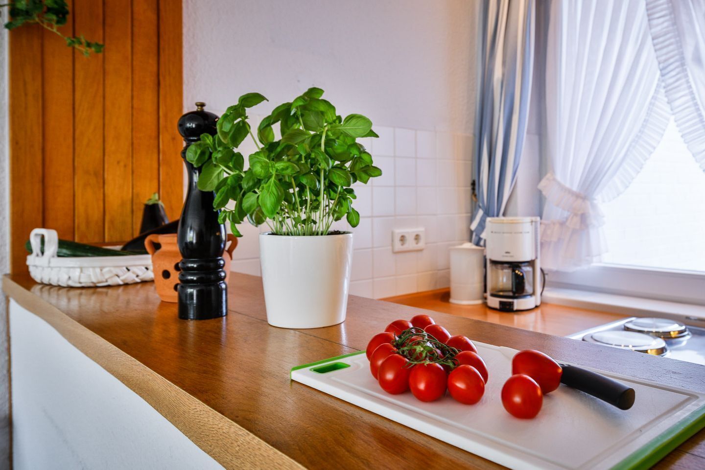 A kitchen counter with a potted basil plant, a pepper grinder, a cutting board with tomatoes, and a coffee maker.