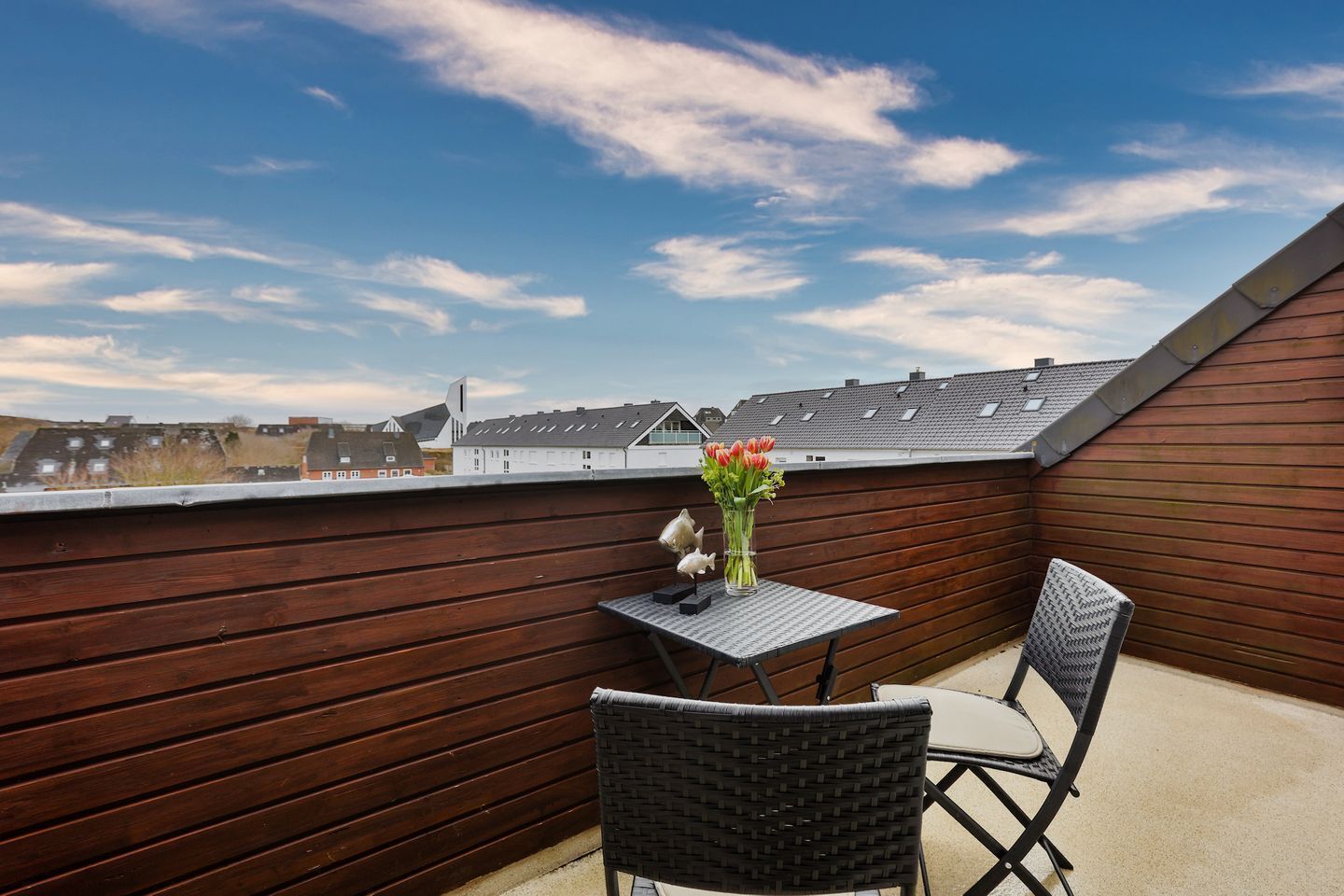 Eine Dachterrasse mit einer Holzverkleidung, einem kleinen Tisch mit Blumen in einer Vase und zwei Korbstühlen.