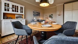 Un comedor con una mesa redonda de madera dispuesta para cuatro personas, rodeada de sillas tapizadas. Una lámpara colgante moderna ilumina la mesa.