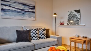 Un salón moderno con un sofá gris, cojines estampados, un frutero sobre una mesa, una lámpara de pie y obras de arte en las paredes.